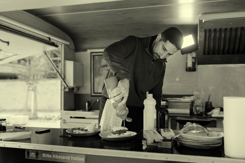 A chef putting some ketchup on a burger for a customer while cooking in their food truck.